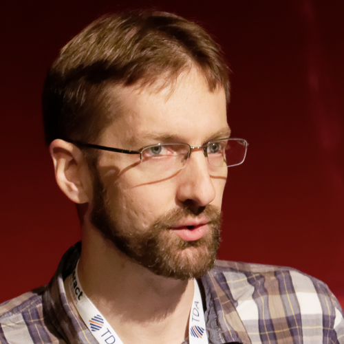 Alastair Campbell profile picture. White male with brown hair and beard, wearing glasses. Dark red background and looking to the right.
